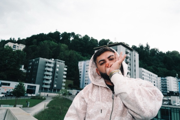 GABU im weissen Kapuzenpullover, Hand am Kinn, vor Hochhäusern